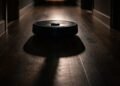 Robot vacuum in a dark hallway casting a long shadow, hinting at a robot vacuum army risk